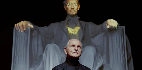 Artist Krysztof Wodiczko in front a statue of Abraham Lincoln, Lincoln is depicted with sunglasses, a necklace and natural hands