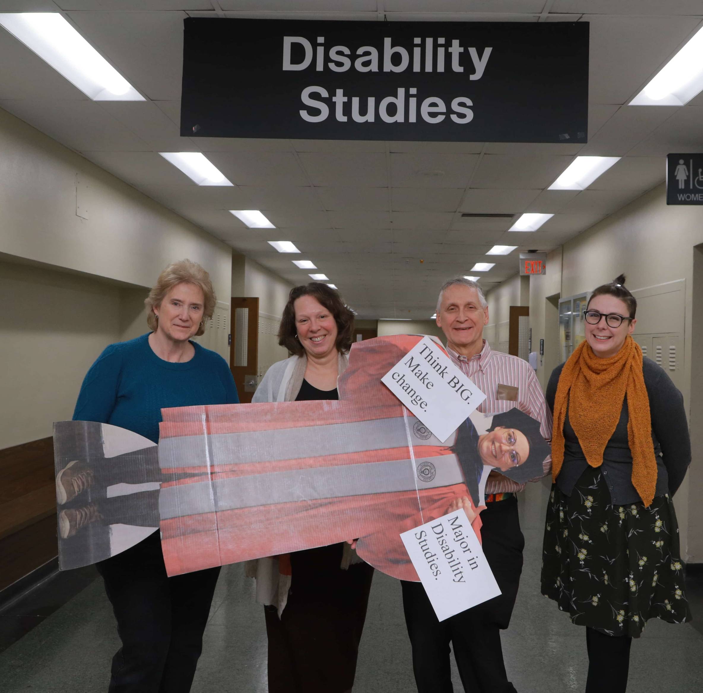 Linda Curtis and professors Kim Nielsen, Jim Ferris, and Becca Monteleone stand smiling under a sign that says Disability Studies while holding a cardboard cutout of professor Ally Day.
