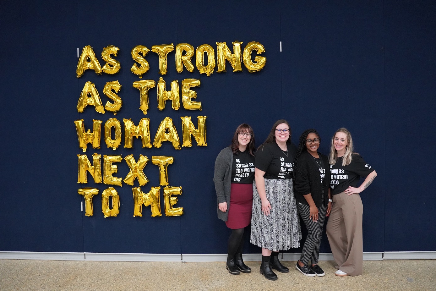 four people next to a foil balloon message that reads, "as strong as the woman next to me"