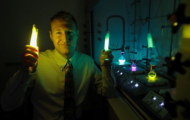 Isaac Schiefer, Ph.D., holding up glowsticks in a darkened laboratory with other glowing chemicals nearby.