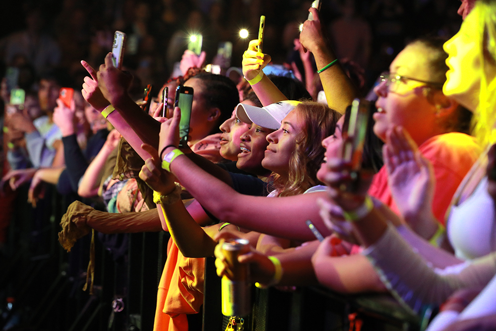 A crowd of smiling spectators watch the concert and record it on their phones