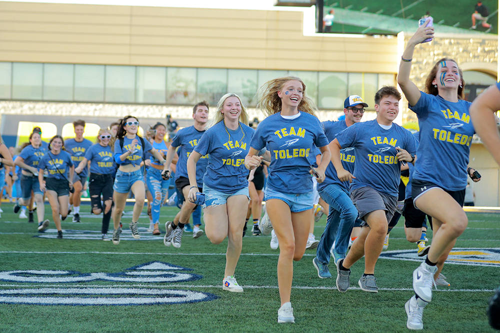 A group of UToledo students run smiling onto the field in the Glass Bowl