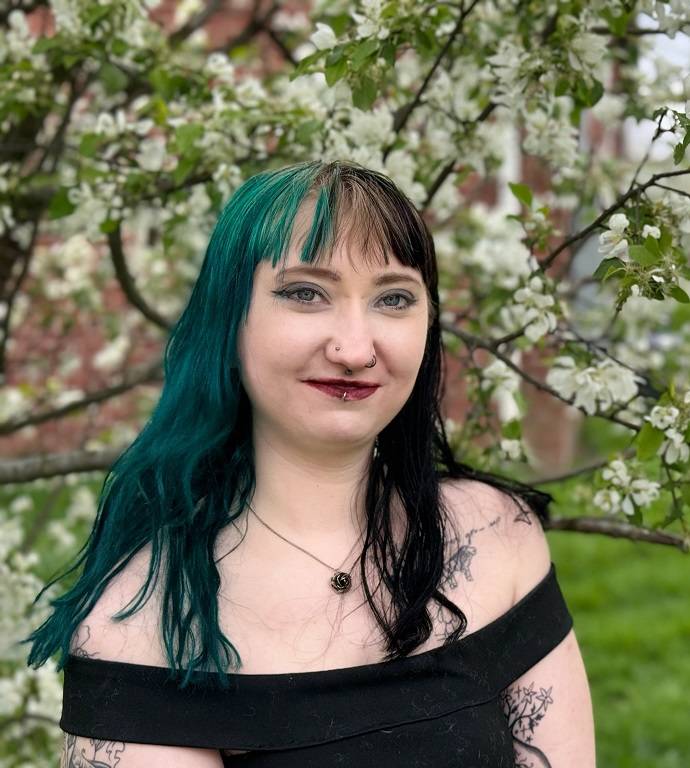 A woman with black and teal hair, Bailey, grinning in front of a blooming tree. 