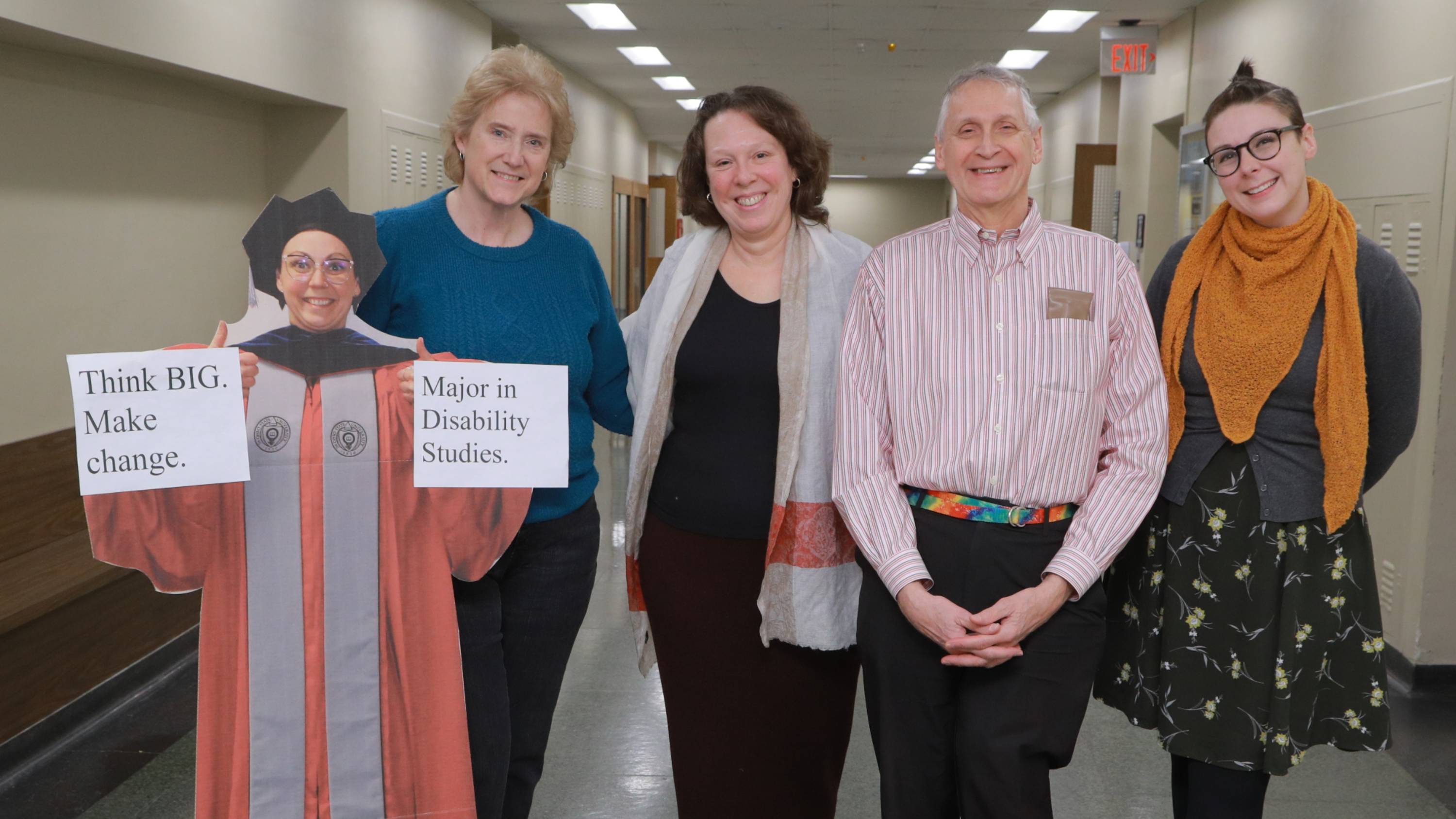Disability Studies office administrator Linda Curtis standing with professors Kim Nielsen, Jim Ferris, and Becca Monteleone, smiling in a hallway. They are next to a cardboard cutout of professor Ally Day with signs that say "Think Big, Make Change. Major in Disability Studies