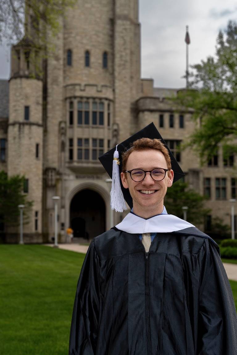 Ryan smiles at the camera in front of University Hall, a sandy stone building with a gothic tower. He is wearing a graduate robe and mortarboard.