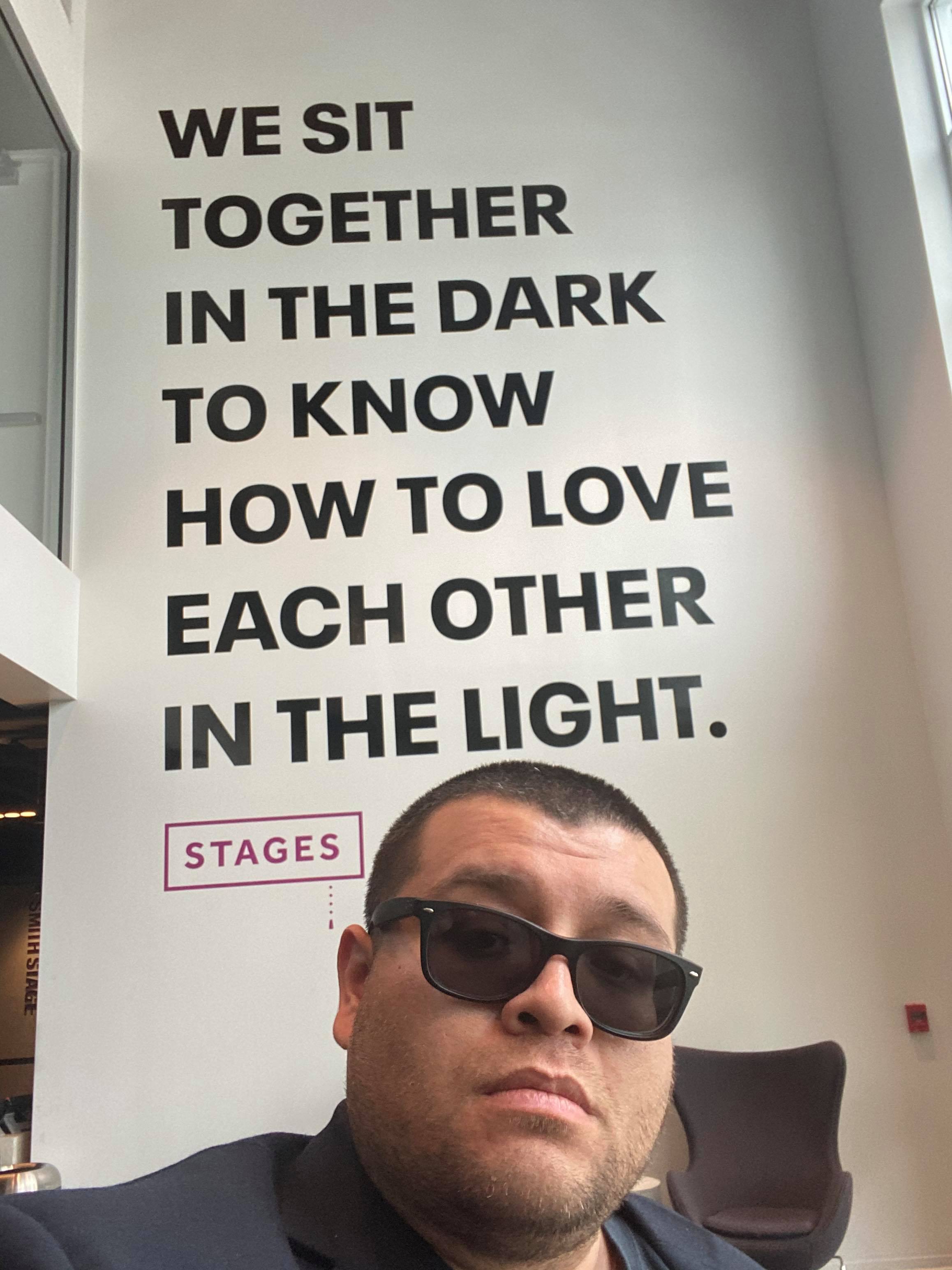 Photo of playwright and tv writer Franky Gonzalez in front of a sign which reads We sit together in the dark to know how to love each other in the light