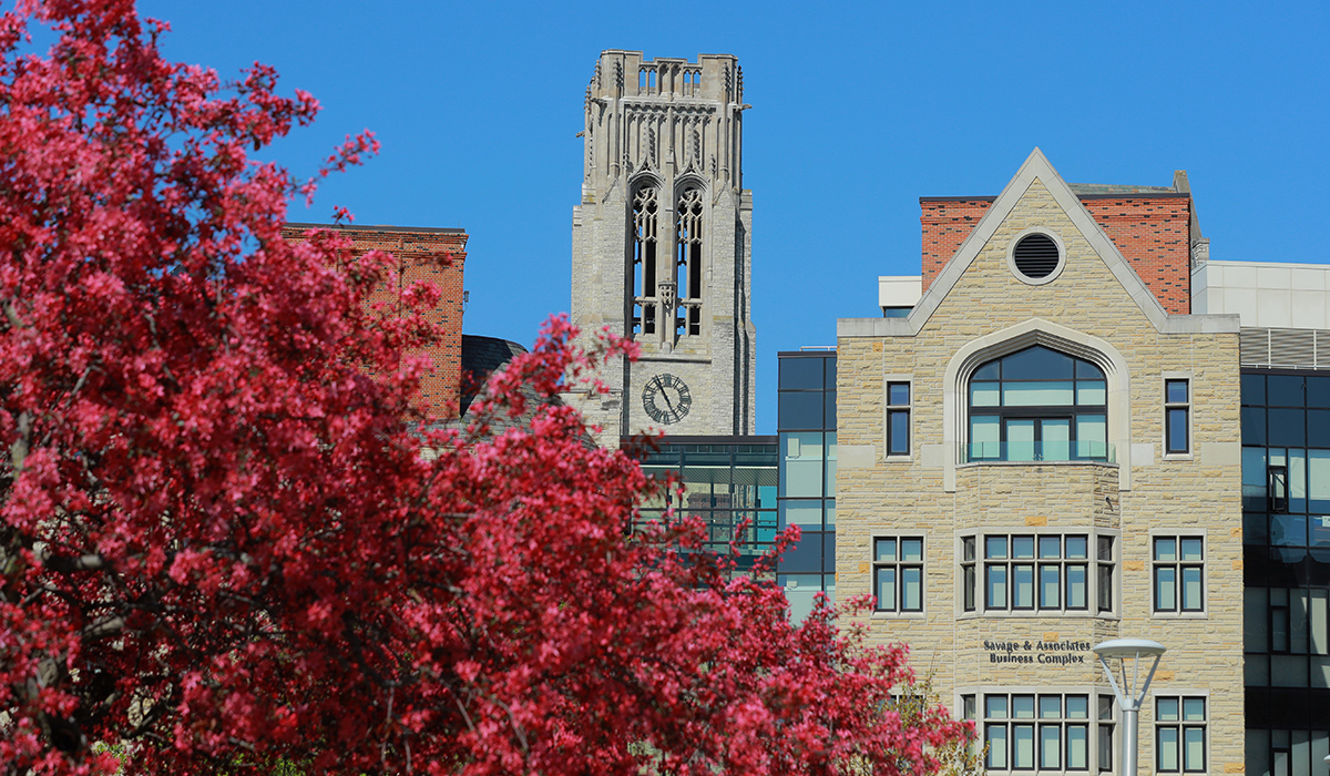 A tree with vibrant red leaves stands in the foreground, with the Savage & Associates Complex and University Hall visible in the background on the University of Toledo campus.