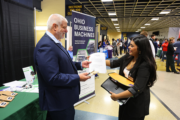 a female college of business student talking with an employer at the business job fair. The businss student is handing over her resume to the employer she is speaking with.