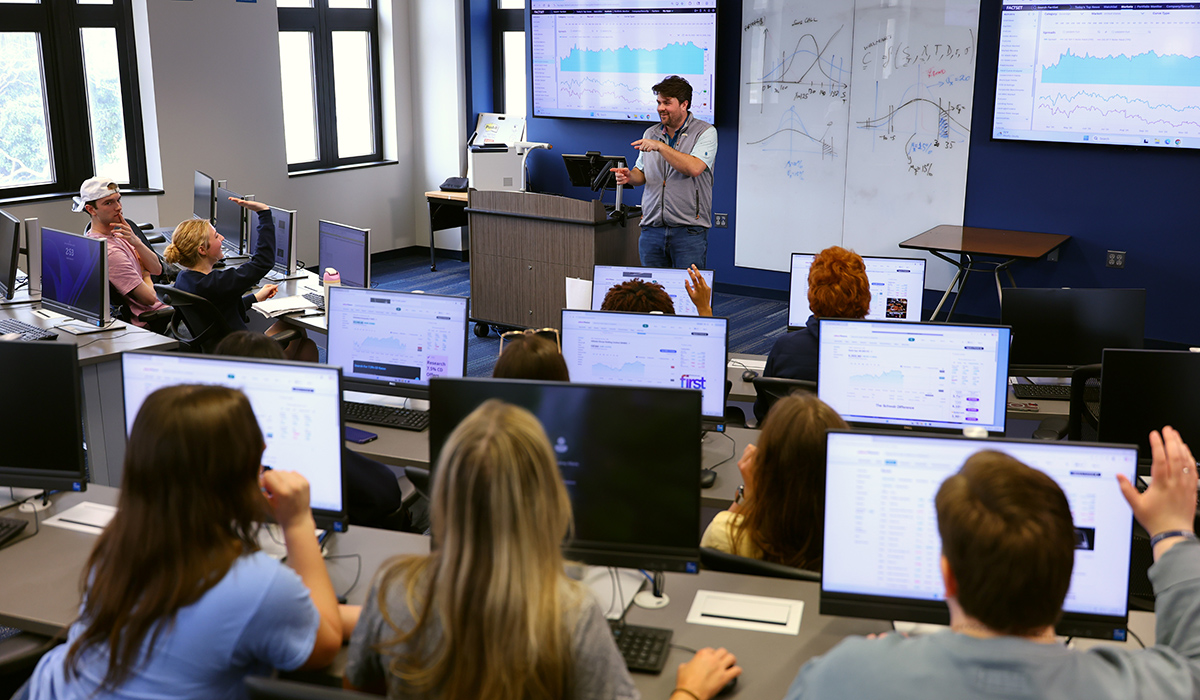 Associate Professor Collin Gilstrap teaching a class in the neff trading room calling on a student with their hand raised