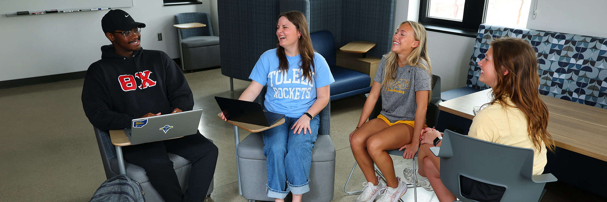 A group of business students sitting on chairs in a class room, working on laptops, talking to each other and laughing
