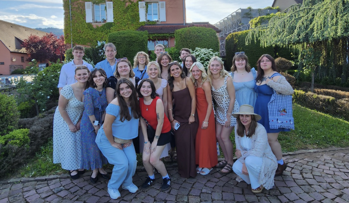 Business students on a study abroad trip standing for a group photo. The group is standing on a brick pathway with moss covered building behind them.
