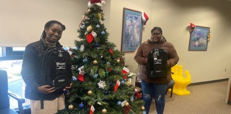 Two members of the Young Executive Scholars pose in front of a holiday tree holding their brand new backpacks.
