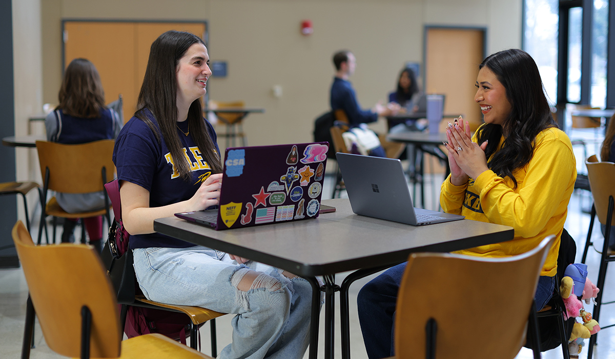 Two college of Business Students sitting at a table with their laptops doing school work and talking to each other