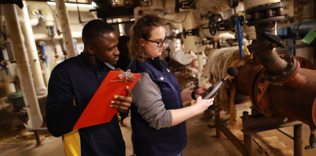 an African American student with a red clipboard and a white student studying pipes and engineering equipment  