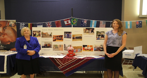 Two females posing for picture in front of a table with a board filled with pictures