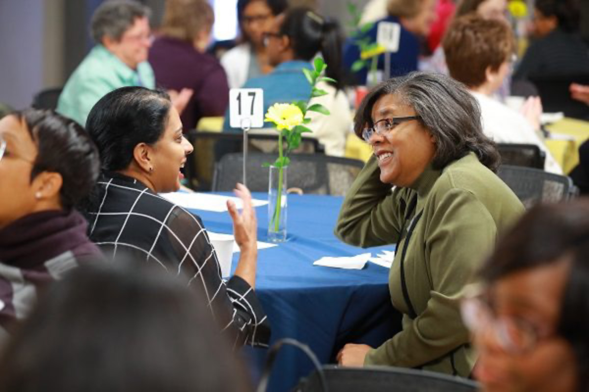 A photo of two female faculty members talking while sitting at a table. 