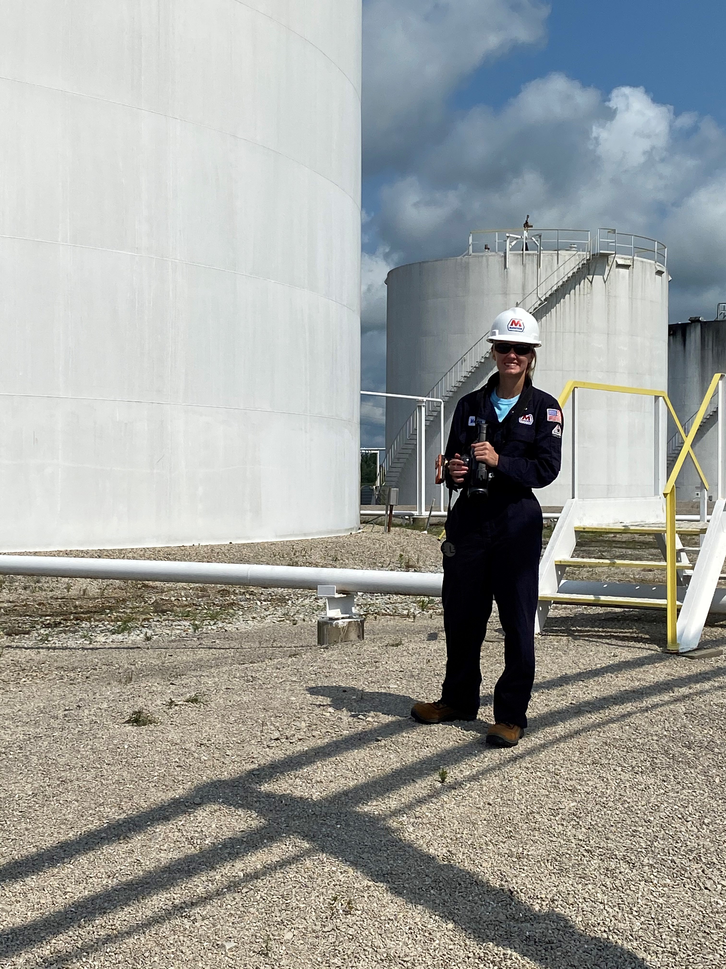 smiling woman in hard hat and blue jumpsuit by oil tank