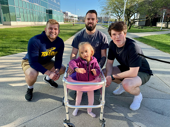 3 male students squatting by little girl in walker