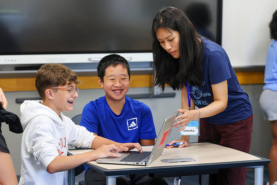 Two boys and a woman work together around an open laptop in a classroom.