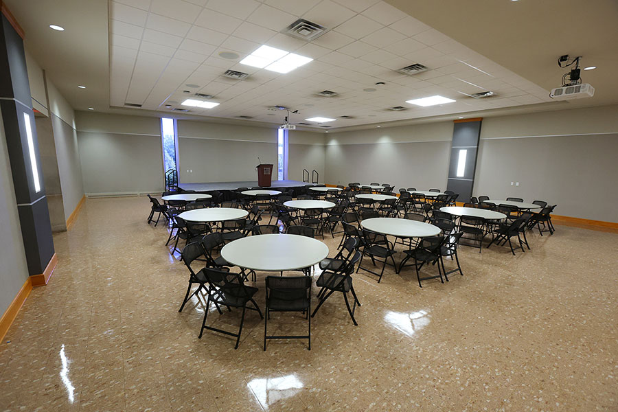 empty large room arranged with round tables, black folding chairs, a small stage, and a podium