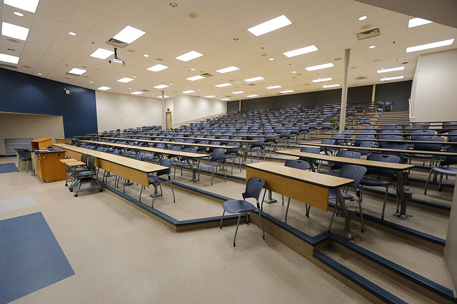 Empty tiered lecture hall with rows of blue chairs and long wooden tables, a lectern, projector, and projection screen.
