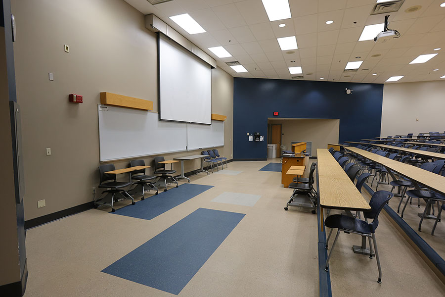 Empty tiered lecture hall with rows of blue chairs and long wooden tables, a lectern, projector, and projection screen.