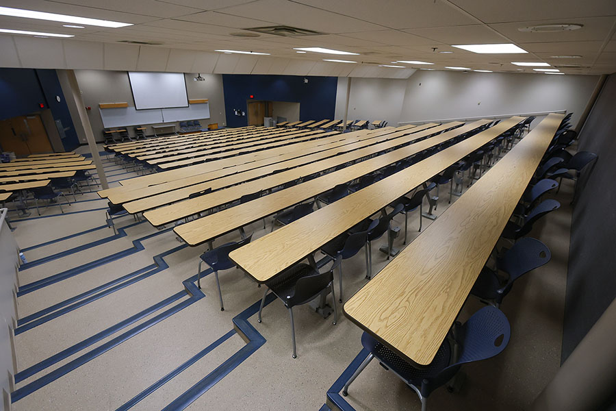 Empty tiered lecture hall with rows of blue chairs and long wooden tables, a lectern, projector, and projection screen.