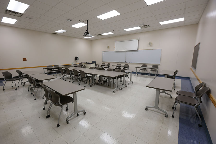 Empty classroom with rows of tables and rolling chairs, a whiteboard, projection screen, and wall-mounted display.