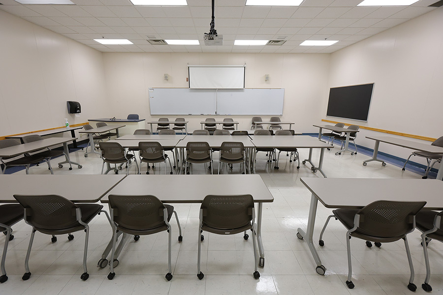 Empty classroom with rows of tables and rolling chairs, a whiteboard, projection screen, and wall-mounted display.