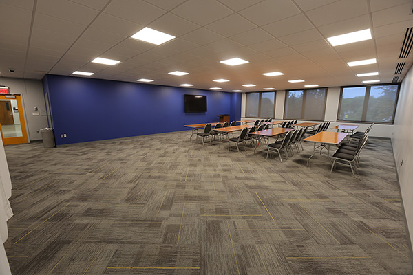 Classroom with rows of wooden tables and black chairs, large windows, and gray carpet