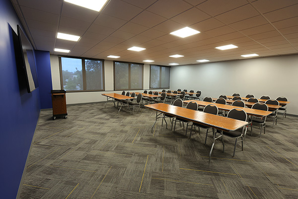 Classroom with rows of wooden tables and black chairs, large windows, and gray carpet