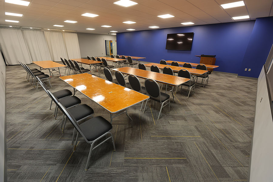 Classroom with rows of wooden tables and black chairs, large windows, and gray carpet