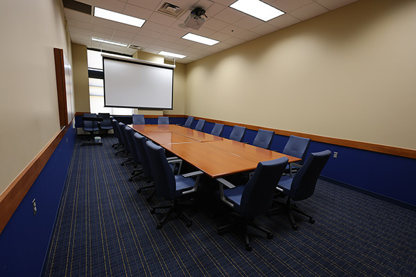 Conference room with a long wooden table, blue chairs, patterned carpet, and a blank projection screen