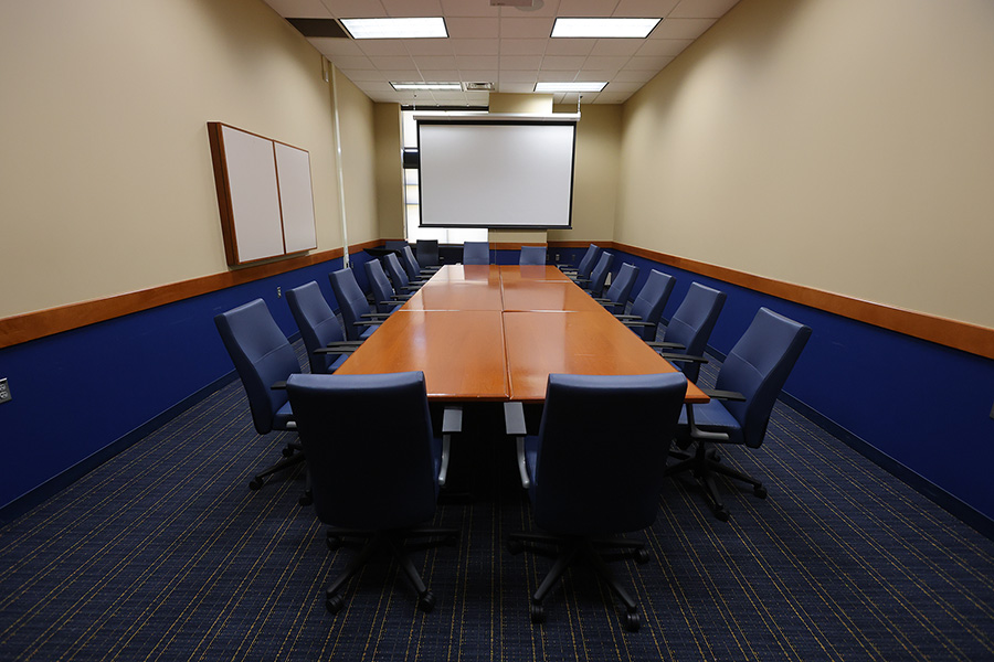 Conference room with a long wooden table, blue chairs, patterned carpet, and a blank projection screen