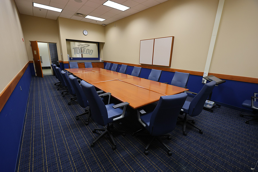 Conference room with a long wooden table, blue chairs, patterned carpet, and a small countertop area with mirror and utoledo rockets logo