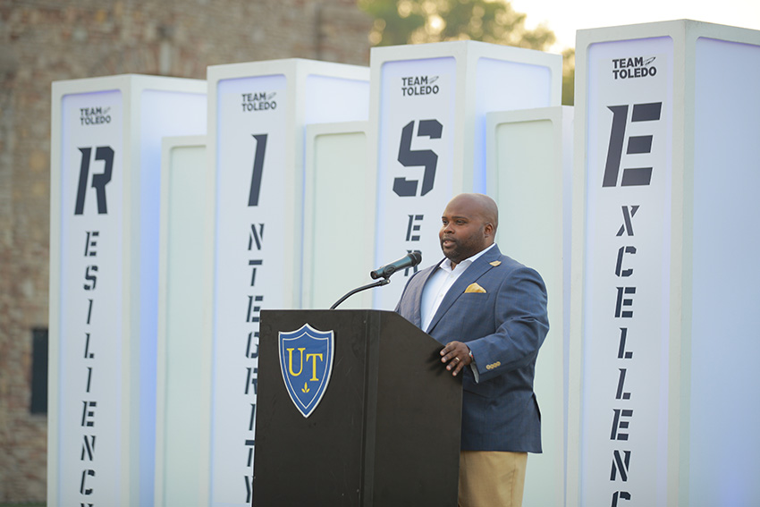 A UToledo representative speaking at a podium in front of four pillars that spell out RISE
