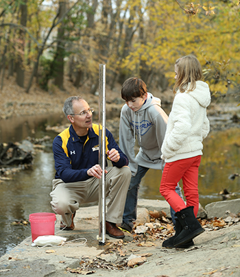 A UToledo professor showing science equipment to a pair of children at the bank of a river.