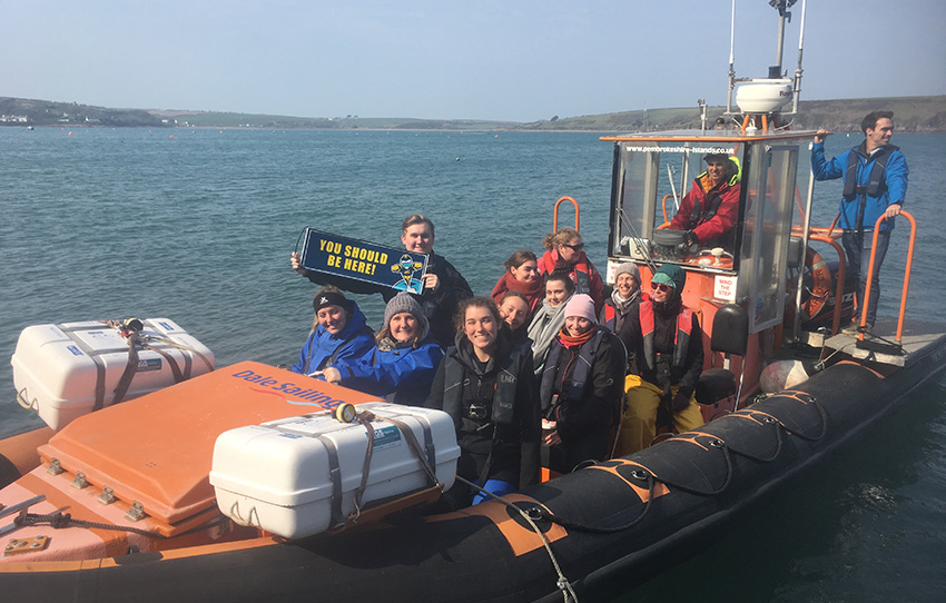 A group of about 10 students gathered on a boat on a body of water. One student is holding a sign that says 'You should be here!'.