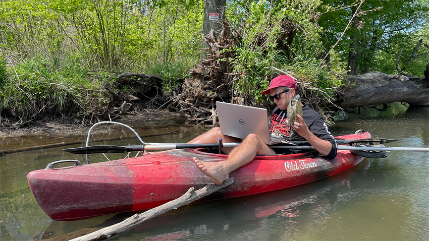 A man sitting in a kayak on a body of water. He is using one hand to hold a turtle and using his other hand to work with a laptop.