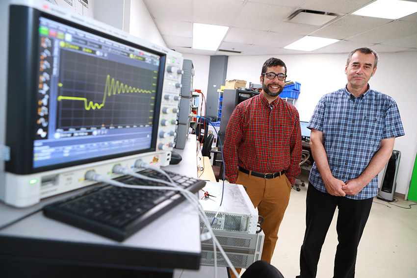 Daniel Georgiev, Ph.D., and Raghav Khanna, Ph.D., standing in a laboratory. A nearby monitor shows a wave pattern.