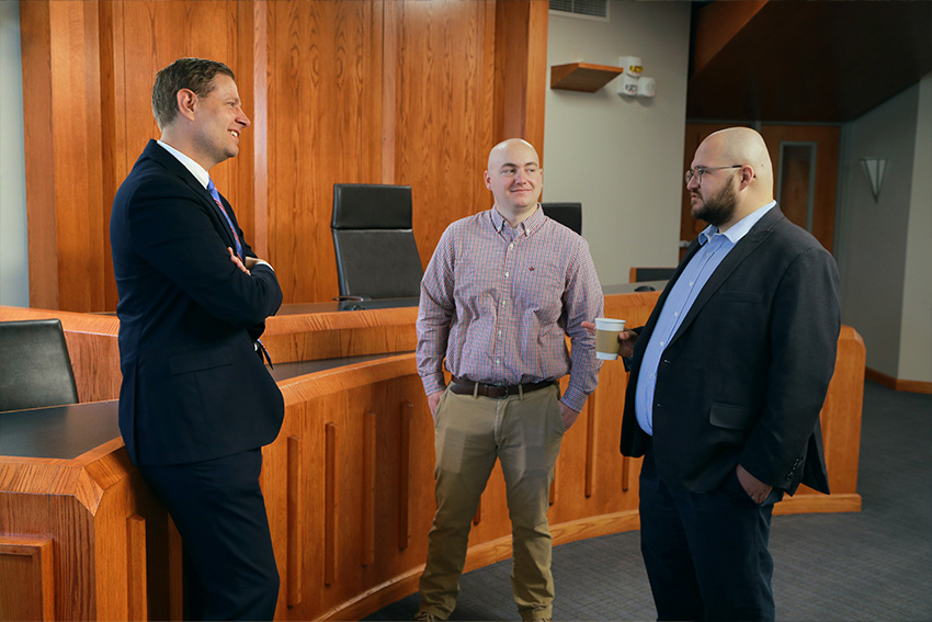 Three professionally dressed men talking in a UToledo courtroom.