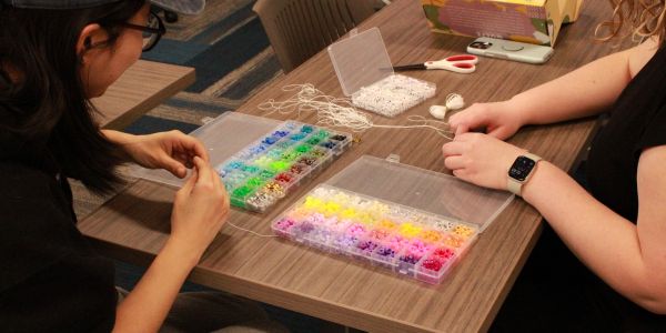 Students seated at table with box of colorful beads and making bracelets