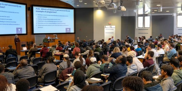 Presenter addressing full lecture hall of students with slide deck.