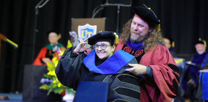 Doctoral student in black regalia and glasses is hooded by advisor in red regalia