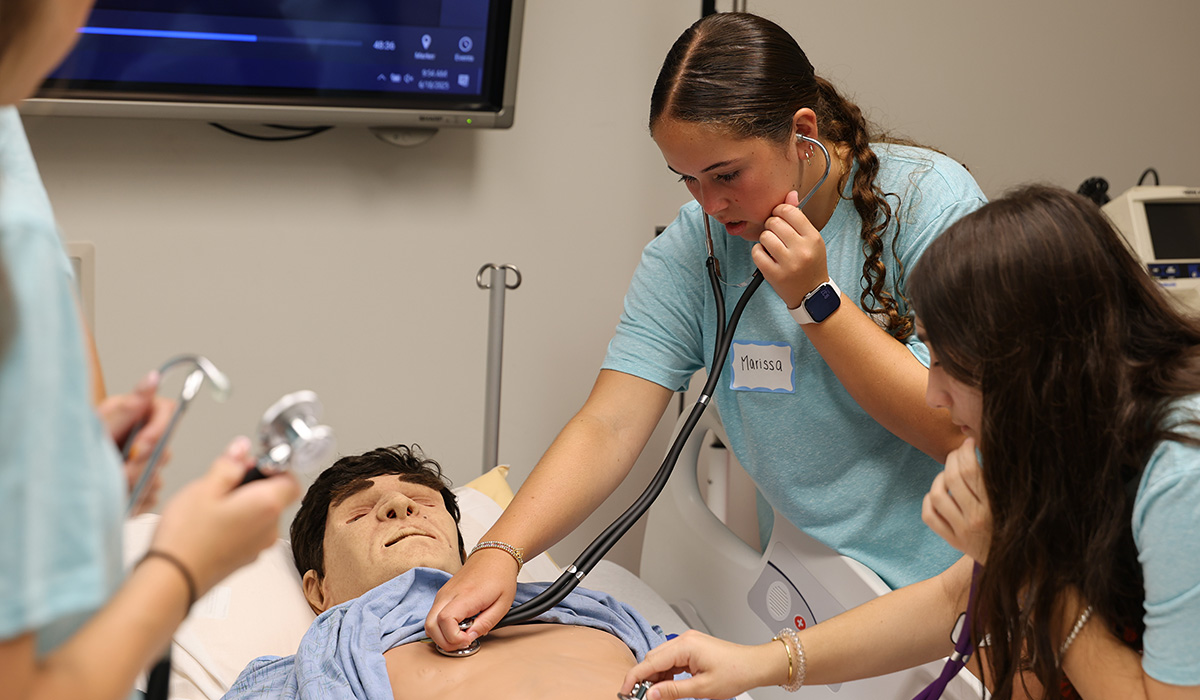 Three women standing around a training mannequin in hospital bed. The women are using stethoscope's on the training mannequin