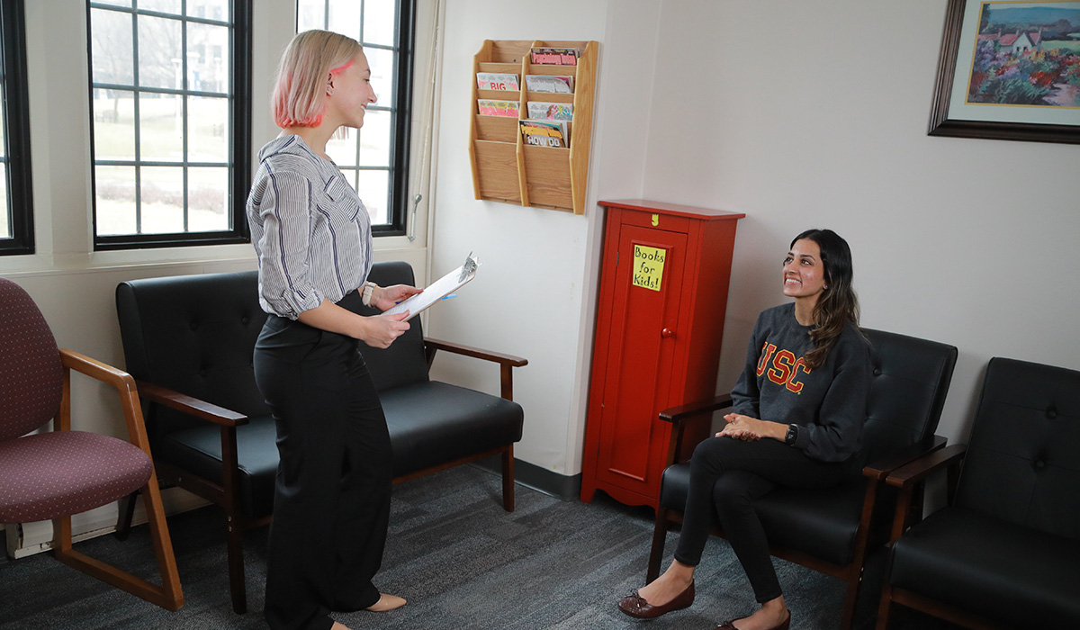 a woman standing holding a clipboard smiles and greets a woman sitting in a chair in a waiting room