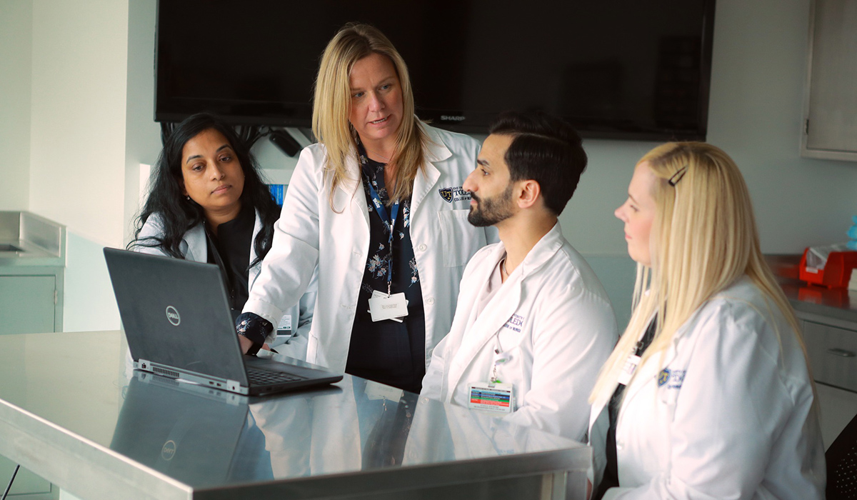 A nursing professor standing at a table in a classroom with a laptop surrounded by 3 nursing students 