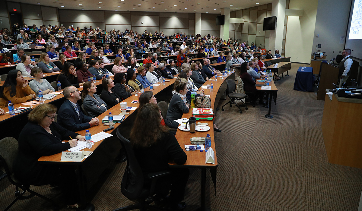 A lecture hall room full of people watching a listening to the speaker giving a presentation