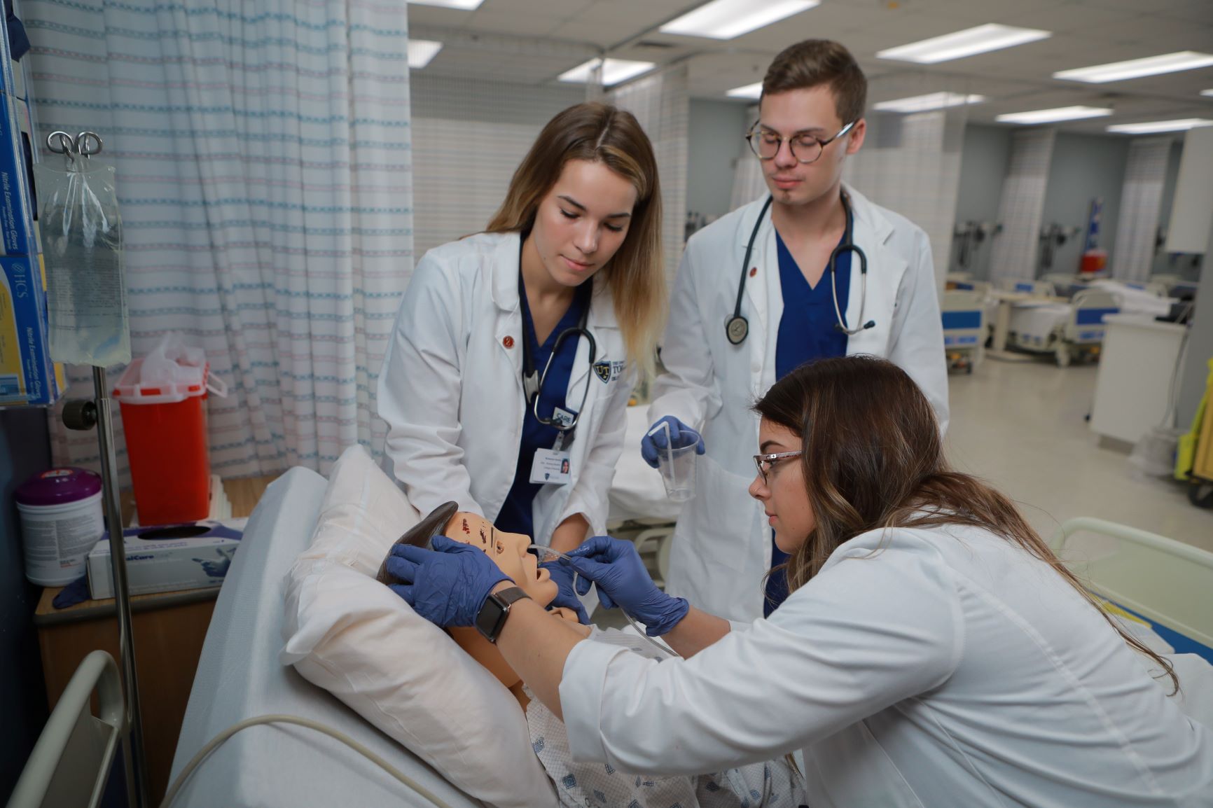 3 nursing students in a medical classroom using medical devices on a testing doll