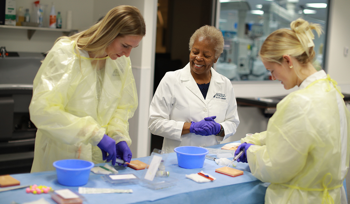 Nursing students and a nursing professor in a classroom standing around a table with medical training equipment